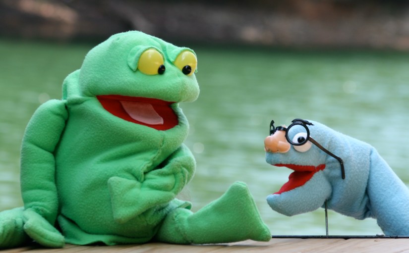 Big green Tiddalick the Frog hand a rod puppet and blue Eel rod puppet posing in front of a lake