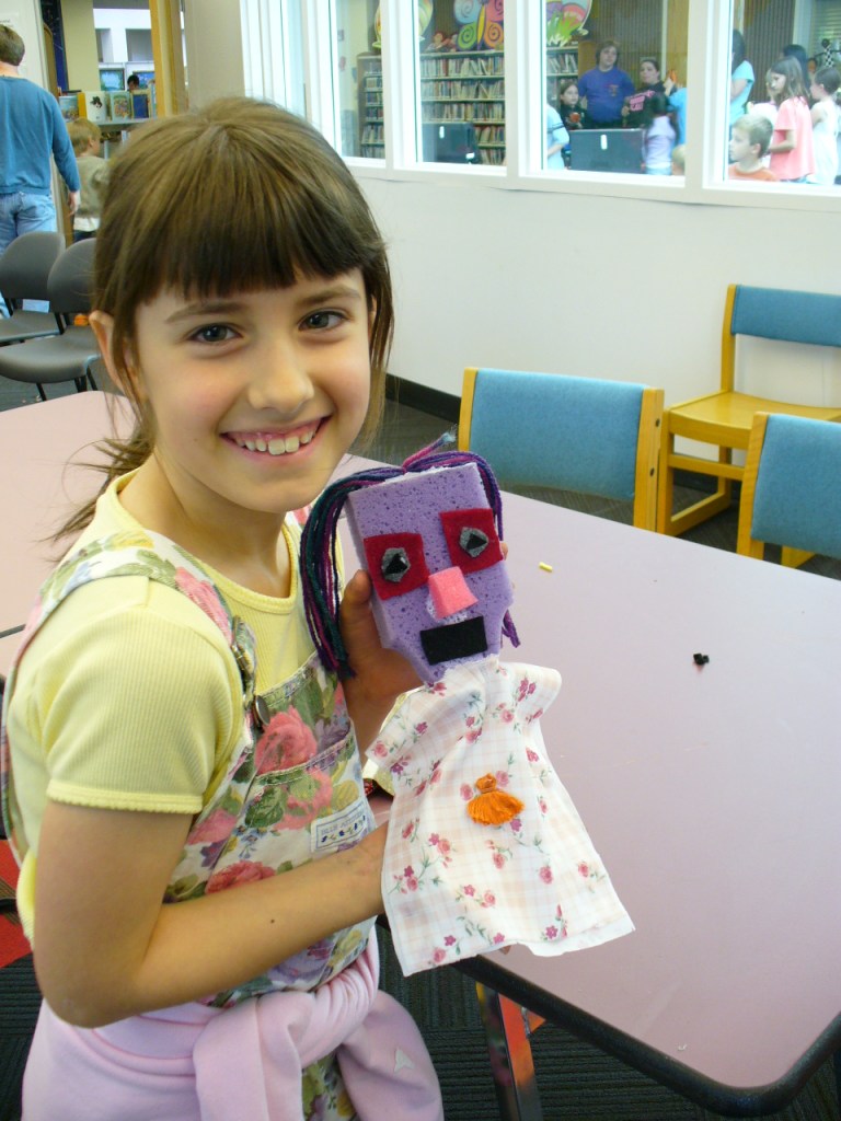 A girl holding a puppet with a purple sponge head and flowered cloth body