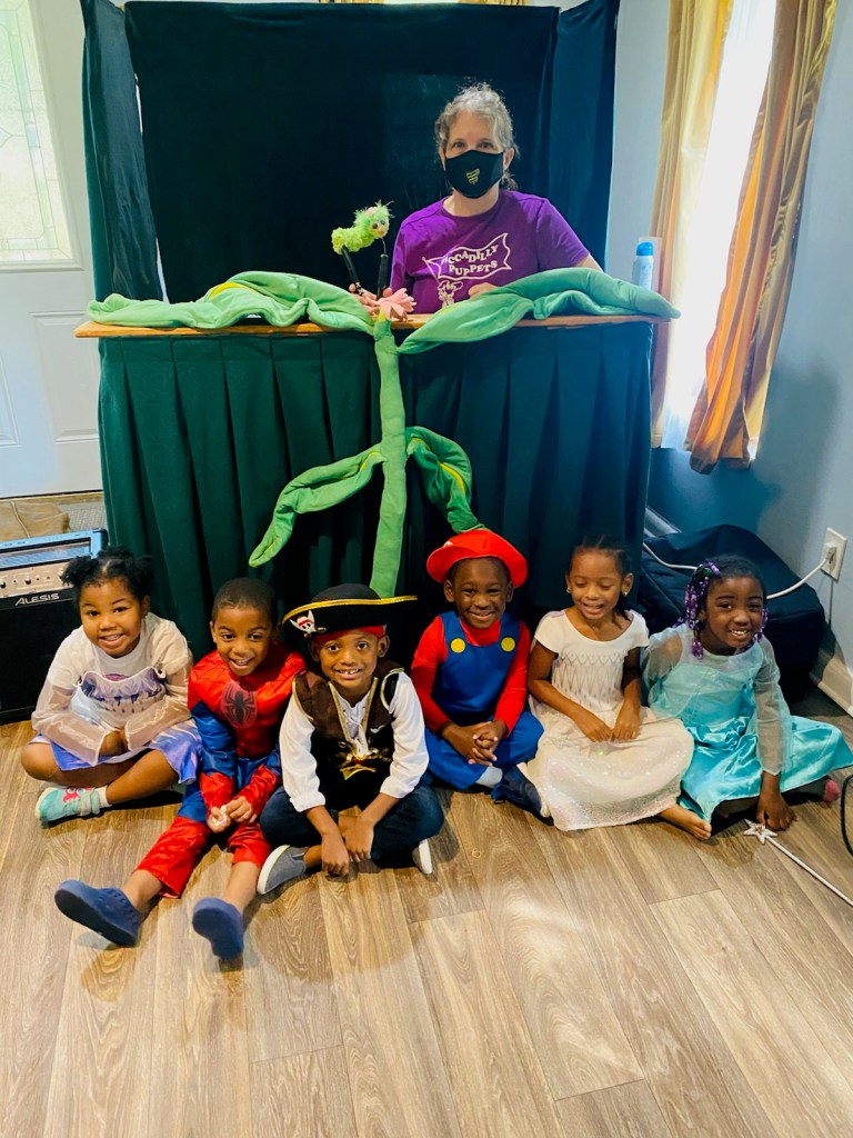 Nancy with small green caterpillar puppet, posing with preschoolers dressed as princesses, Spiderman and a pirate in a house.